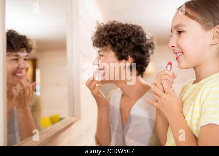 Fröhliche kleine Mädchen Anwendung roten Lippenstift, während ihre Mutter setzen Lippenbalsam Stockfoto