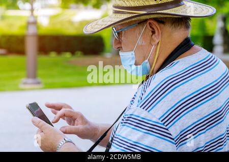 Porträt eines alten Mannes eine medizinische Maske mit Verwendung Smartphone beim Hören Schützen Sie während des Coronavirus COVID-19 in einem Park Stockfoto