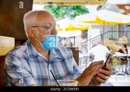 Alter Mann mit Smartphone, weiß eine tragende medizinische Maske schützen während Coronavirus COVID-19 in einem Park Stockfoto