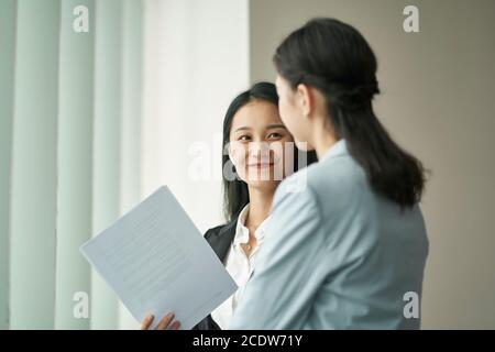 Zwei junge asiatische Geschäftsfrauen stehen am Fenster mit einem Diskussion im Büro Stockfoto