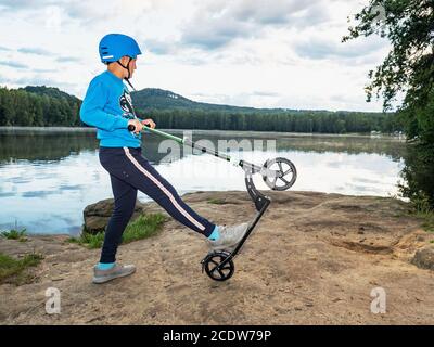 Junge mit Roller macht Trick auf felsigen Seeufer vor dem Hintergrund des Parks. Stockfoto