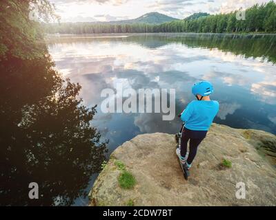 Junge mit Roller macht Trick auf felsigen Seeufer vor dem Hintergrund des Parks. Stockfoto