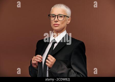 Junge elegante blonde Frau in schwarzem Anzug, Krawatte und Brille, die Sie anschaut Stockfoto
