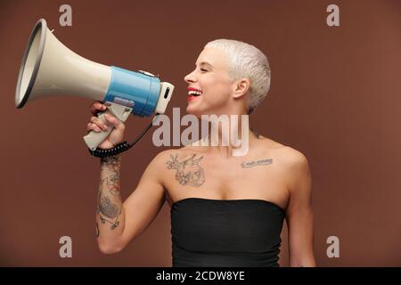 Glückliche junge blonde Frau mit Tattoos auf Arm, Brust und Hals hält Megaphon Stockfoto