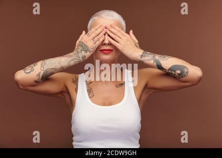 Maskuline Frau in der Weste, die die Augen von den Händen bedeckt und zeigt Arme mit Tattoos Stockfoto