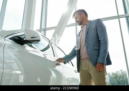 Moderner junger Mann in smarter Casualwear, der durch neues Weiß steht Auto Stockfoto