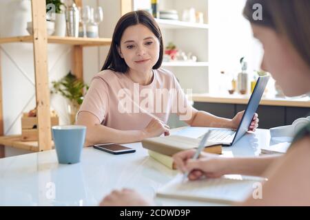 Junge Mutter mit Laptop, die ihre Tochter anschaut, die Notizen macht Im Copybook Stockfoto