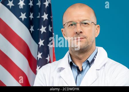 Glatzköpfiger Arzt mittleren Alters in Brillen und Weißkopfflächen, die dagegen stehen US-Flagge Stockfoto
