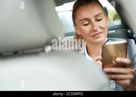 Auf dem Weg zum Geschäftstreffen. Porträt einer schönen und erfolgreichen Geschäftsfrau mit ihrem Smartphone und lächelnd, während sie auf dem Rücksitz sitzt Stockfoto