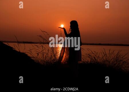 Am 5. Januar 2020 orange Sonnenuntergang hinter Gras Silhouette Mädchen Frau hält Sonne auf der Hand in Bakerswar Damm, birbhum, West Bengal, Indien Stockfoto