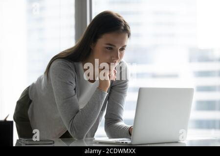 Fokussierte junge Geschäftsfrau, die im Büro am Tisch steht und einen Laptop benutzt Stockfoto