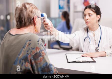 Junge Arzt mit Infrarot-Thermometer, um ältere Frau Temperatur im Krankenhaus zu nehmen. Medic trägt weißen Mantel und Stethoskop und Krankenschwester am Computer im Hintergrund. Stockfoto
