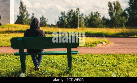 Der einsame Mann sitzt auf einer Bank in einem Kapuzenpullover mit Blick auf die grüne Parklandlandschaft voller Bäume und Gras. Rückansicht. Kreative Outd Stockfoto