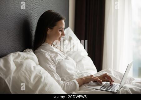 Lächelnde junge Frau im Bademantel mit Laptop, sitzend im Bett Stockfoto