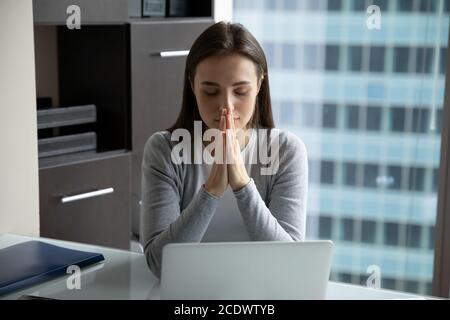Hoffnungsvolle Wunschfrau junge Geschäftsfrau, die betet, am Schreibtisch im Büro sitzt Stockfoto