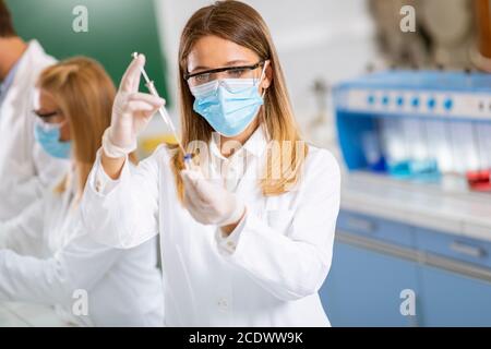 Frau Arzt trägt schützende Gesichtsmaske und Sicherheit googles in Labornadelspritze und Impfstoffflasche für Medikamentenfläschchen Stockfoto