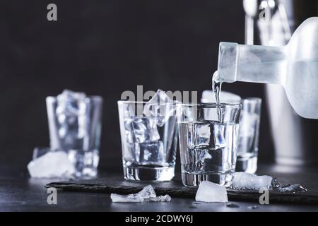 Wodka in Schnapsgläsern, die aus der Flasche auf schwarzem Stein-Hintergrund ausgießen, eisgekühltes starkes Getränk in Nebelglas Stockfoto