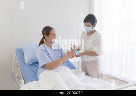 Eine junge asiatische Frau, die in einem Krankenhaus krank war, lag auf einem Erholungsbett. Ihre Mutter, die sich um ihre Seite kümmerte, benutzte Alkoholgel, um ihr ha zu waschen Stockfoto