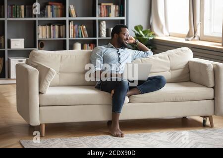 Nachdenklicher afroamerikanischer Mann, der sich auf der Couch mit Laptop entspannt und visualisiert Stockfoto