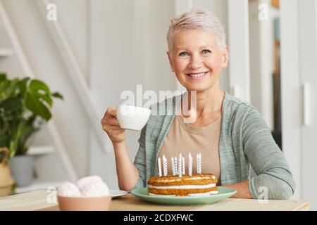 Glückliche ältere Frau lächelt an der Kamera, während sie an der sitzt Tisch und Tee trinken mit Kuchen Stockfoto