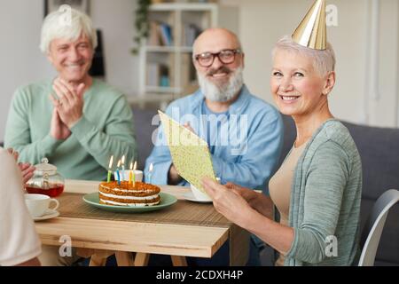 Porträt einer glücklichen älteren Frau, die Grußkarte liest und lächelt Bei der Kamera, während sie mit ihren Freunden Geburtstag feiern Stockfoto