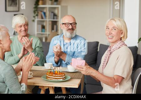 Porträt einer älteren Frau, die Geschenkbox hält und lächelt Kamera mit ihren Freunden sitzen am Tisch mit Kuchen Und applaudieren Stockfoto