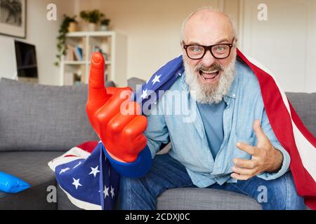 Aufgeregt bärtigen Mann in Brillen mit amerikanischen Flagge sitzen auf Sofa und Jubel für seine Fußballmannschaft zu Hause Stockfoto