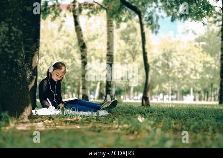 Glückliches Mädchen in weißen Kopfhörern mit digitalen Tablet-pc im Park. Fernlernkonzept. Resilienz, zurück zur Schule, neue Normalität. Stockfoto