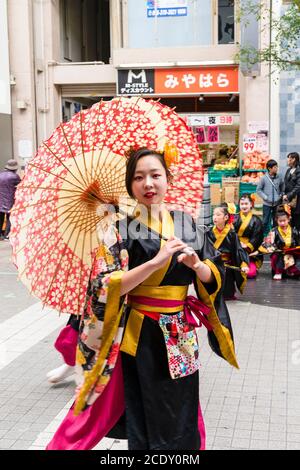 Nahaufnahme einer japanischen Frau, die Teil eines yosakoi-Tanzteams ist, die tanzt, während sie einen Sonnenschirm hält und direkt auf den Zuschauer schaut. Kyusyu Gassai Festival. Stockfoto