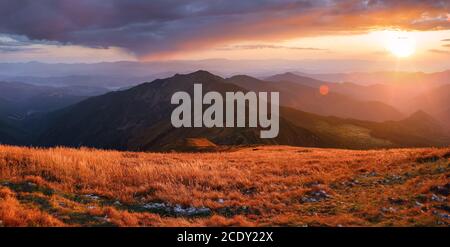 Herbstlandschaft. Erstaunlicher Sonnenuntergang erhellt die Umgebung. Vom Rasen im orangefarbenen Gras, ein Panoramablick auf den hohen Berg, Himmel mit Wolken. Stockfoto