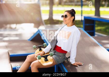 Junge Frau mit einem Longboard in den Händen ruht Im Park zum Skateboarden an einem sonnigen Sommermorgen Stockfoto