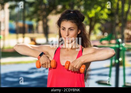 Junge Frau führt Übungen mit Hanteln auf dem Spielplatz an einem warmen sonnigen Morgen. Gesundes Lifestyle-Konzept Stockfoto