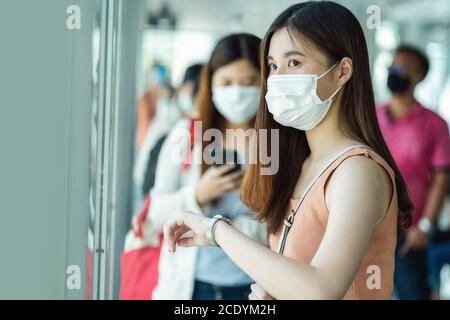 Junge asiatische Frau Passagier trägt chirurgische Maske und Überprüfung der Zeit beim Warten Bus am Bahnhof mit gekrönten Menschen, wenn Reisen in der Großstadt bei Stockfoto