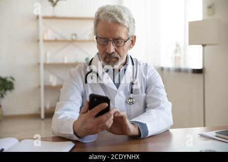 Serious reifen Arzt Blick auf Smartphone-Bildschirm, sitzen am Schreibtisch Stockfoto