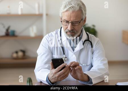 Serious reifen Arzt Blick auf Smartphone-Bildschirm, sitzen am Schreibtisch Stockfoto