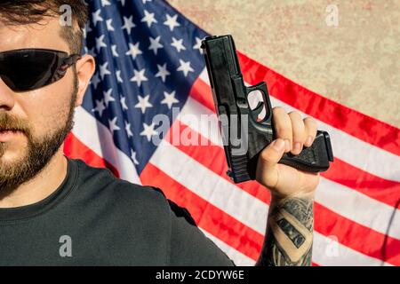 Nahaufnahme eines tapferen jungen bärtigen Mannes mit Sonnenbrille und Pistole Gegen amerikanische Flagge Stockfoto