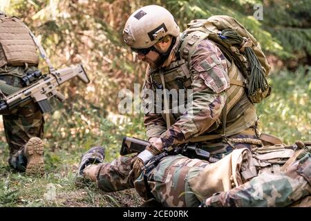 Mann in Helm und Sonnenbrille Untersuchung Bein des verwundeten Soldaten Auf dem Schlachtfeld Stockfoto