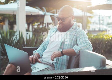 Schöner bärtiger Hipster Mann mit Laptop in einem Sommer-Café im Freien. Remote-Arbeiten von zu Hause oder Café-Konzept. Stockfoto