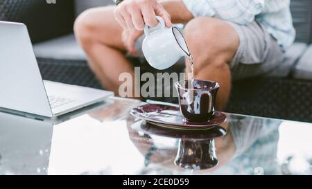 Zugeschnittenes Bild eines jungen Mannes, der Kaffee oder Tee trinkt (heißes Wasser in eine Tasse gießt), der morgens einen Laptop benutzt. Intelligentes Arbeiten von zu Hause oder Café-Konzept Stockfoto