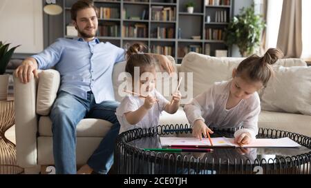 Kleine Schwestern zeichnen Bilder, während der Vater auf dem Sofa ruht. Stockfoto