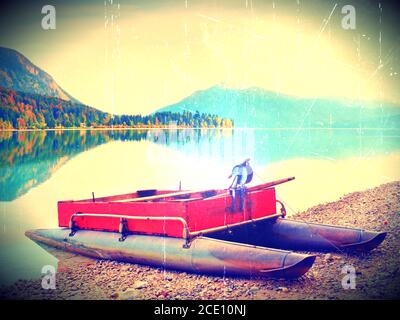 Filmeffekt. Verlassene rot grau Fischerboot am Ufer des Alpensees. Herbstlicher Morgen am See in sanfter Sonneneinstrahlung. Stockfoto