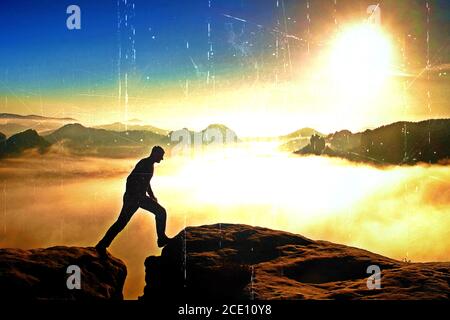 Filmkörnung. Wanderer in Schwarz springt zwischen den felsigen Gipfeln. Wunderbarer Tagesanbruch in felsigen Bergen, schwerer oranger Nebel in der Tiefe Stockfoto