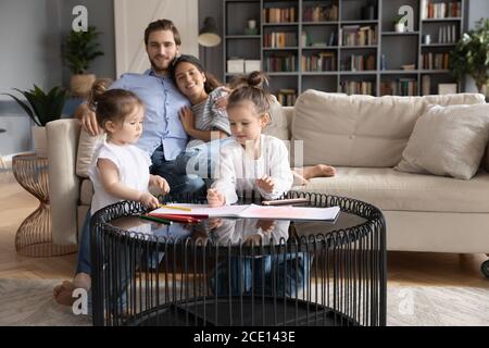 Glücklich liebende junge Paar beobachten zwei kleine Töchter zeichnen Bilder. Stockfoto