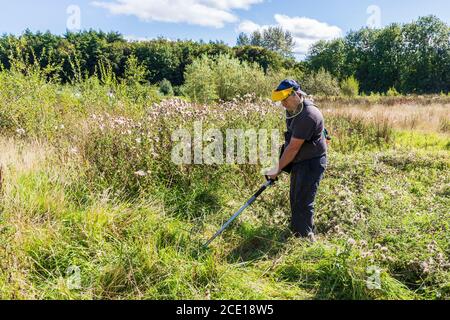 Mann, der einen überwucherten Garten streift, einen industriellen Strimer verwendet und ein Schutzvisier trägt, Kilwinning, Ayrshire, Schottland, Großbritannien Stockfoto