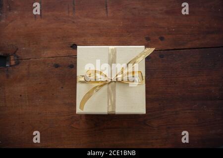 Überraschung mit anwesenden Konzept. Beige geschlossene Geschenkbox Verpackung mit Gold Farbband gegen braunen Holztisch Hintergrund. Draufsicht. Stockfoto