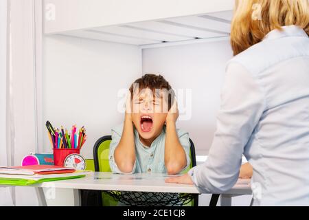 Autistischer Junge schreit und schließt die Ohren bei Schmerzen während der ABA Entwicklungstherapie sitzt am Tisch mit Lehrer Stockfoto