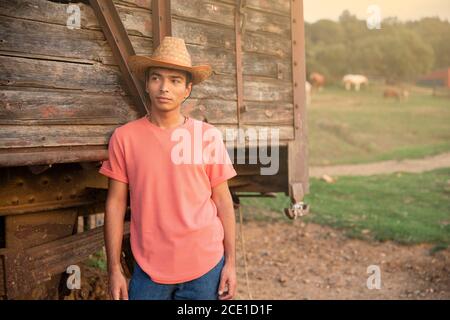 Porträt eines jungen Mannes mit Jeans und einem Cowboy Hut vor einem hölzernen Wagen in der Ranch. Ranch Konzept Fotografie Stockfoto