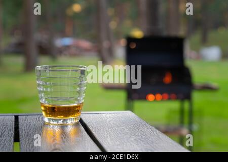 Whiskey Glas auf dem Holztisch neben dem brennenden Grill im Freien im Hinterhof. Alkoholgetränk auf der Terrasse. Niemand Stockfoto