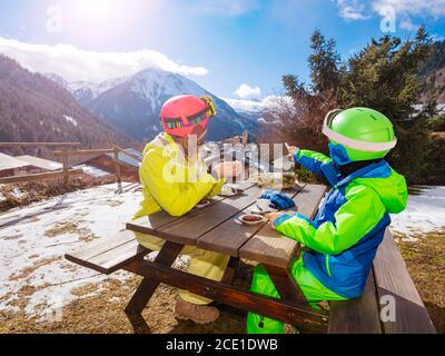 Kleiner Junge im Skioutfit sitzen genießen Mittagspause mit Mutter zeigt mit dem Finger und diskutiert unten das Dorf Stockfoto