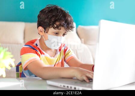 Kleiner Junge mit einem Laptop, um von zu Hause aus zu lernen. Er trägt eine Gesichtsmaske. Konzept der Online-Bildung. Stockfoto